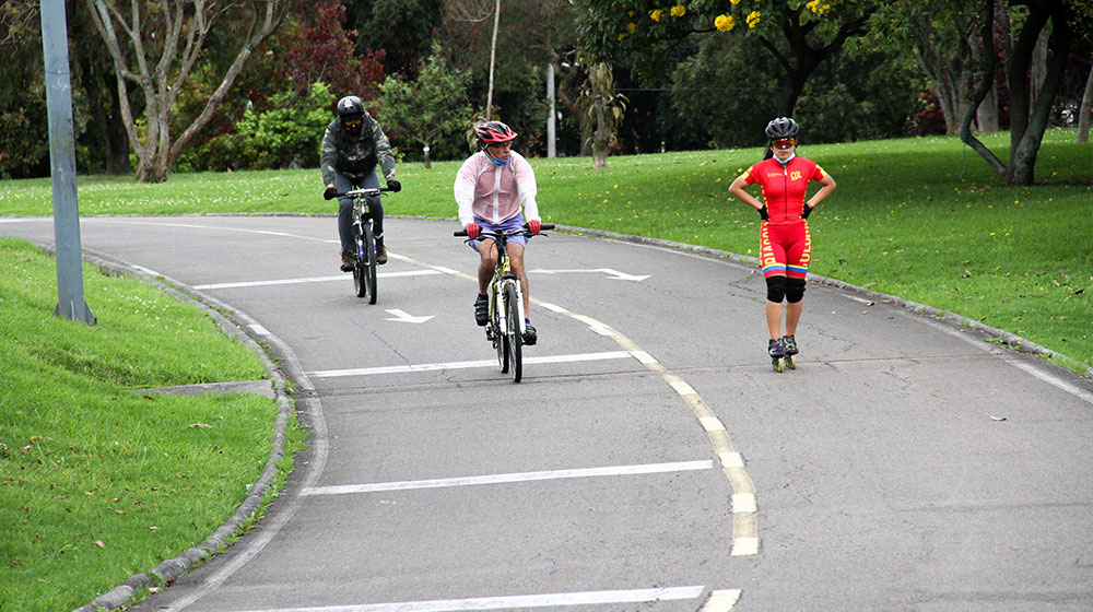 Dos hombres en bicicleta y una joven en patines realizando ejercicio en los alrededores de la Biblioteca Pública Virgilio Barco.