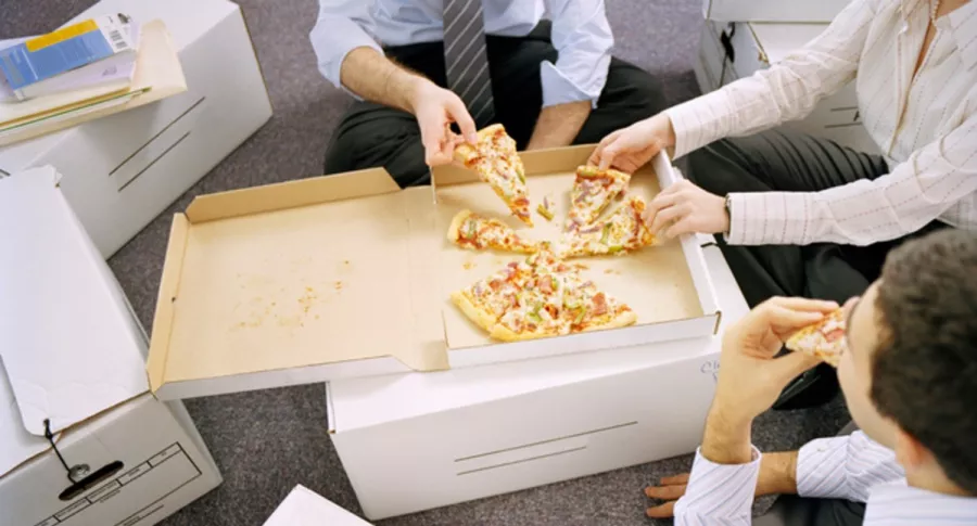 Trabajadores comen pizza en oficina, ilustra nota de millonaria indemnización para mujer que demandó a jefes por no invitarla a comer pizza