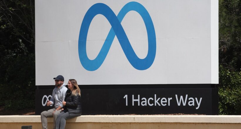 Imagen de dos personas sentadas frente a logo de Meta ilustra artículo Facebook, Amazon, Uber y otras frenan contratación de personal de personal