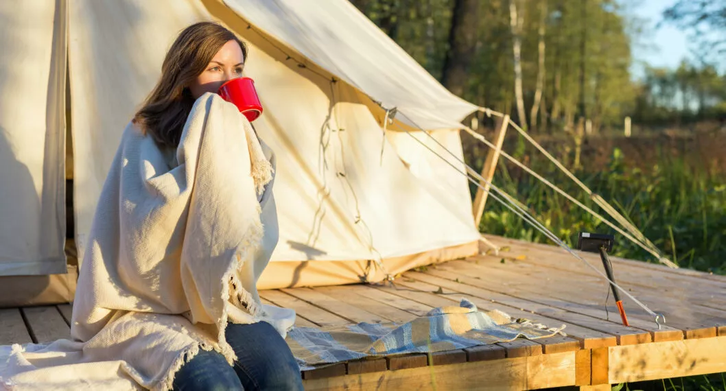 Young woman wraps blanket over herself while sitting and drinking coffee near canvas tent in the morning in the woods