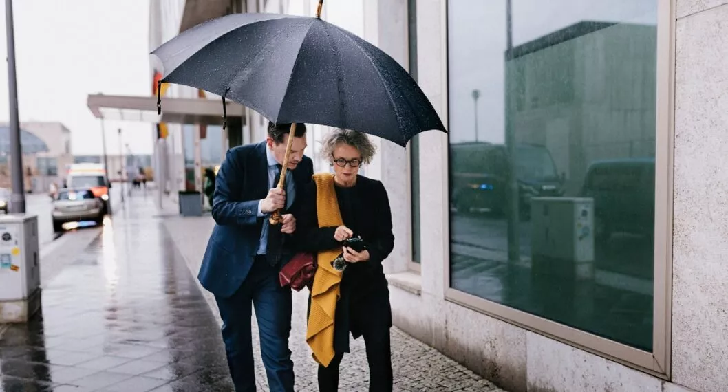 Full body shot of two business people outdoors on a rainy day.  Both are talking together while looking at smartphone.