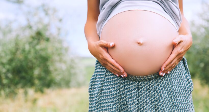 Close-up of pregnant belly in nature, outdoors, copy space. Pregnant woman holds hands on belly on natural background of green grass. Pregnancy, expectation, birth of new life concepts.