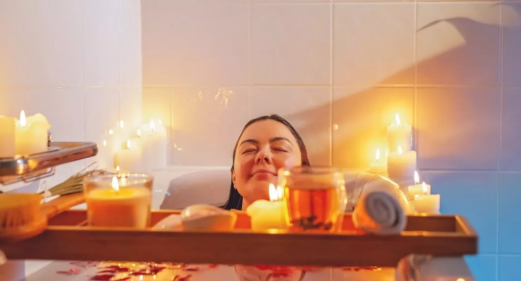 Young woman enjoying spiritual aura cleansing rose flower bath with rose petals and candles during full moon ritual. Body care and mental health routine.