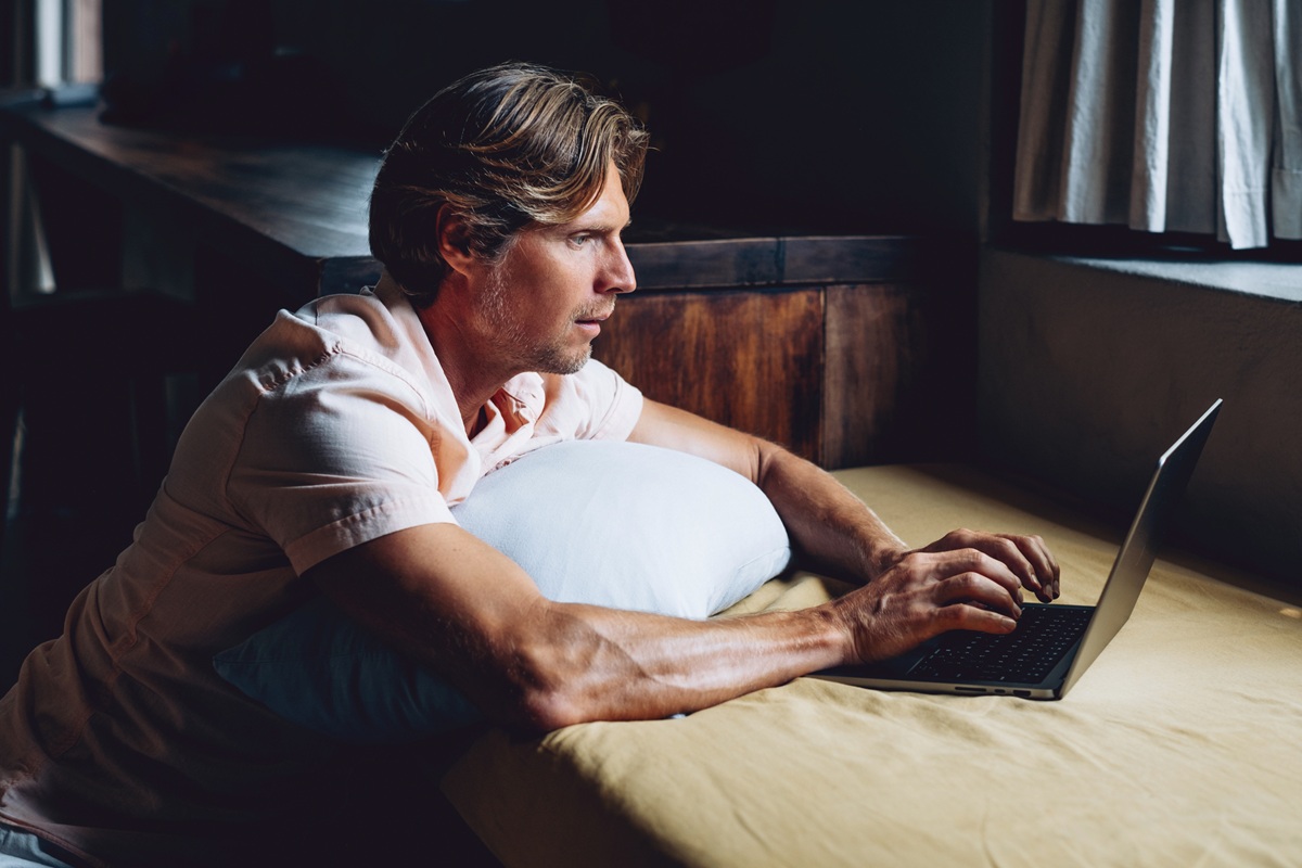 Hombre concentrado usando una computadora portátil en un dormitorio oscuro y acogedor con luz natural de la ventana. Concepto de trabajo remoto y estilo de vida digital moderno.