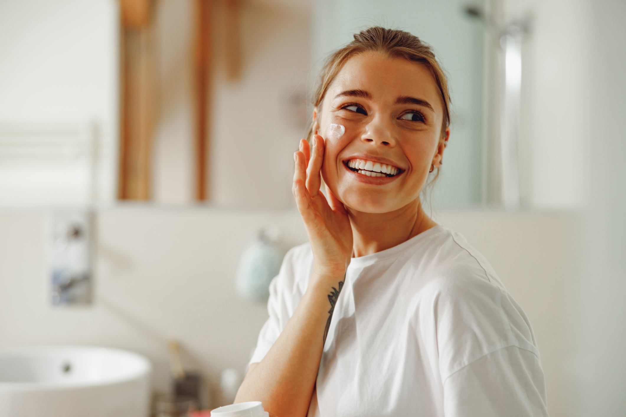 Happy woman applying hydrating moisturizer on her face sitting in bathroom. Home beauty routine