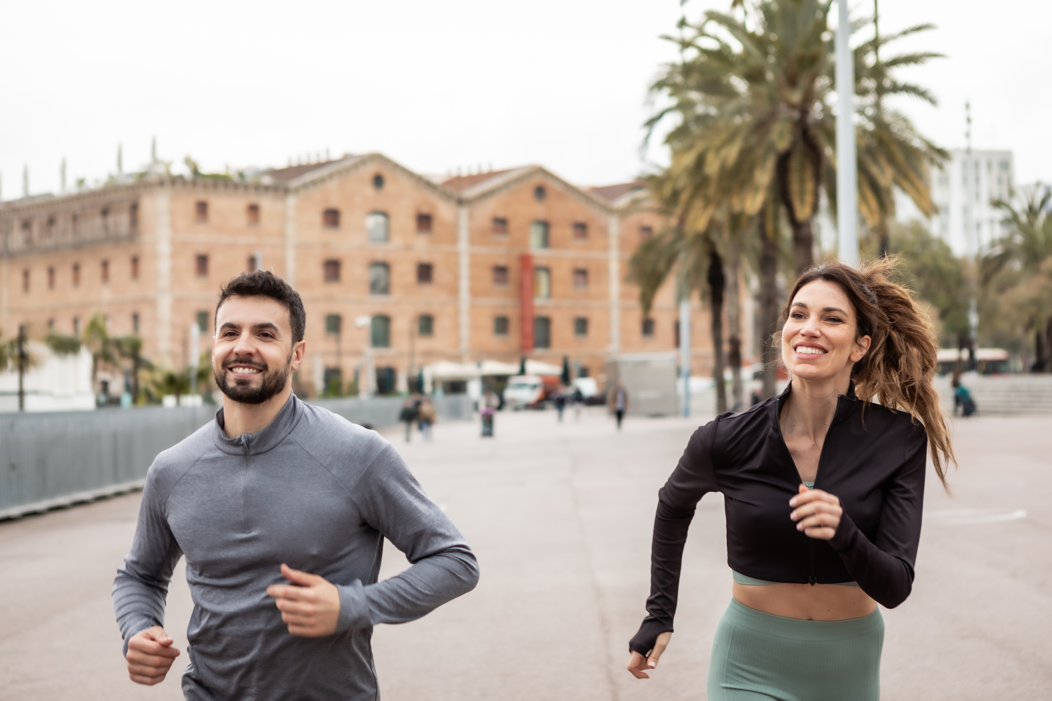 Smiling man and woman participating in a fitness activity, jogging in an urban environment with buildings and palm trees in the background during daytime