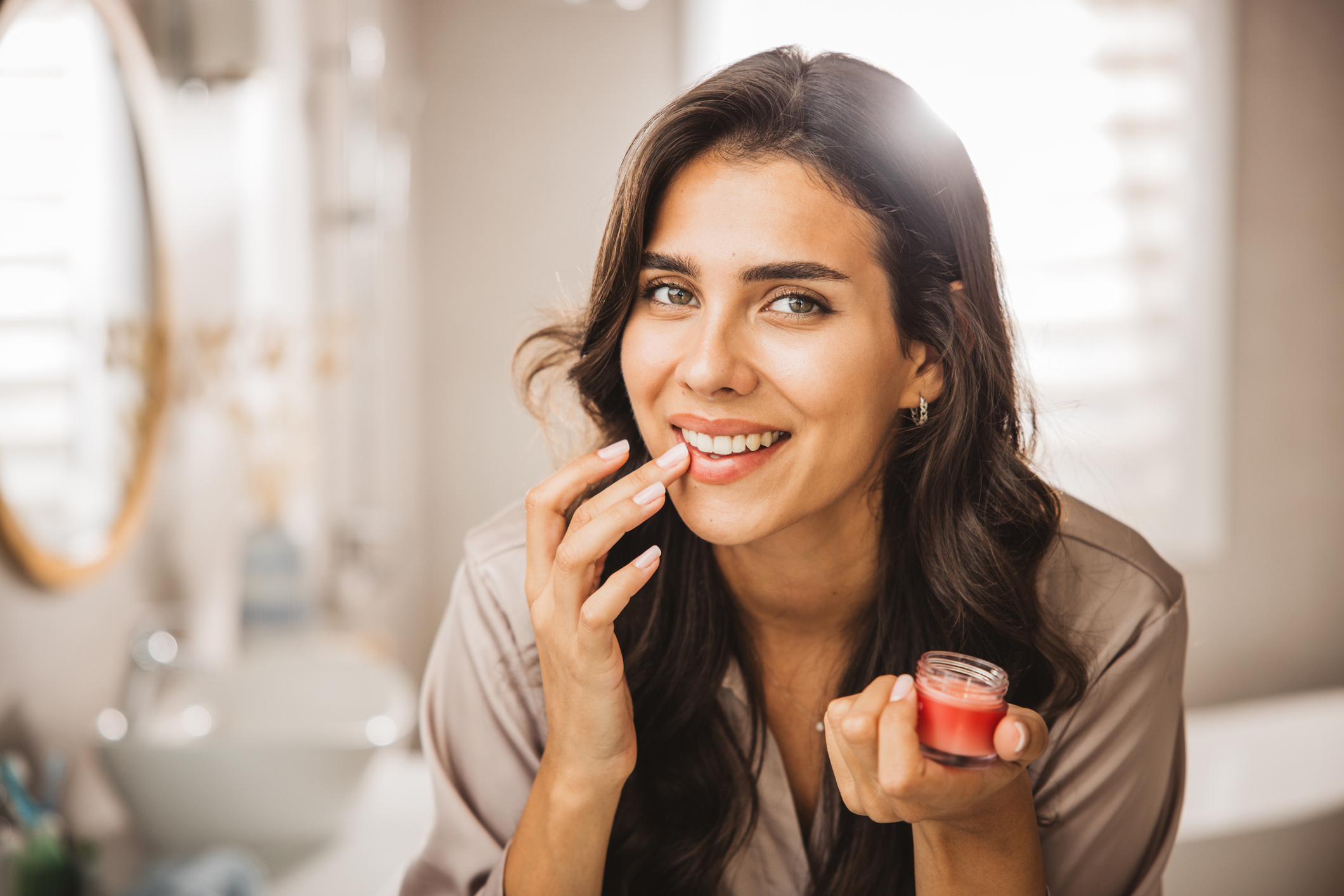 Happy young woman in pajama applying lip gloss in front of mirror in bathroom.