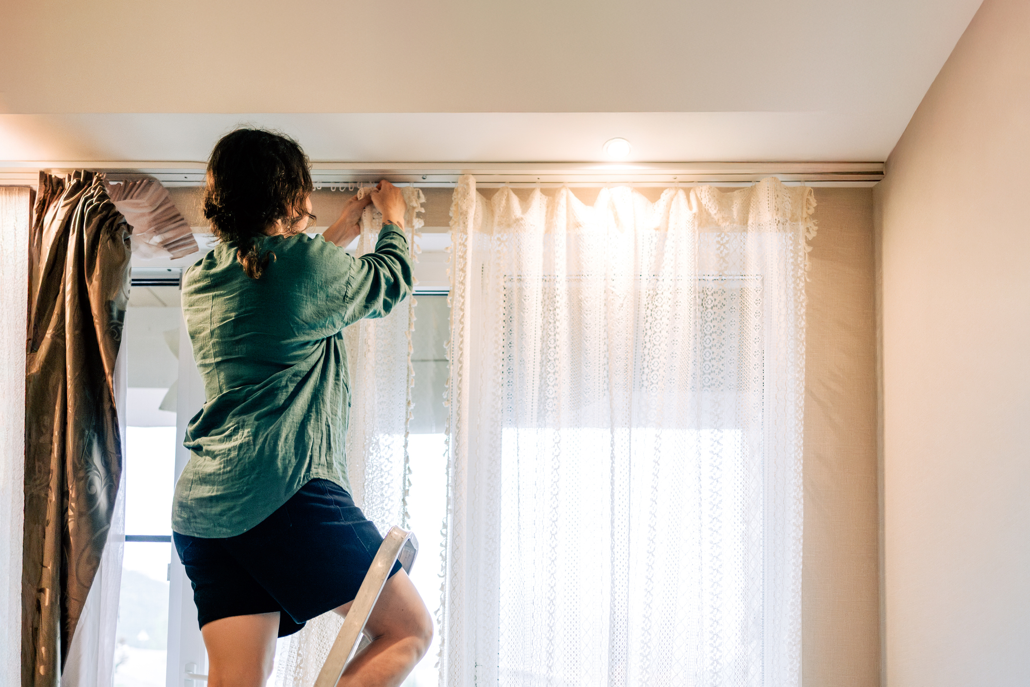 Woman standing on stepladder and hanging curtains in bedroom