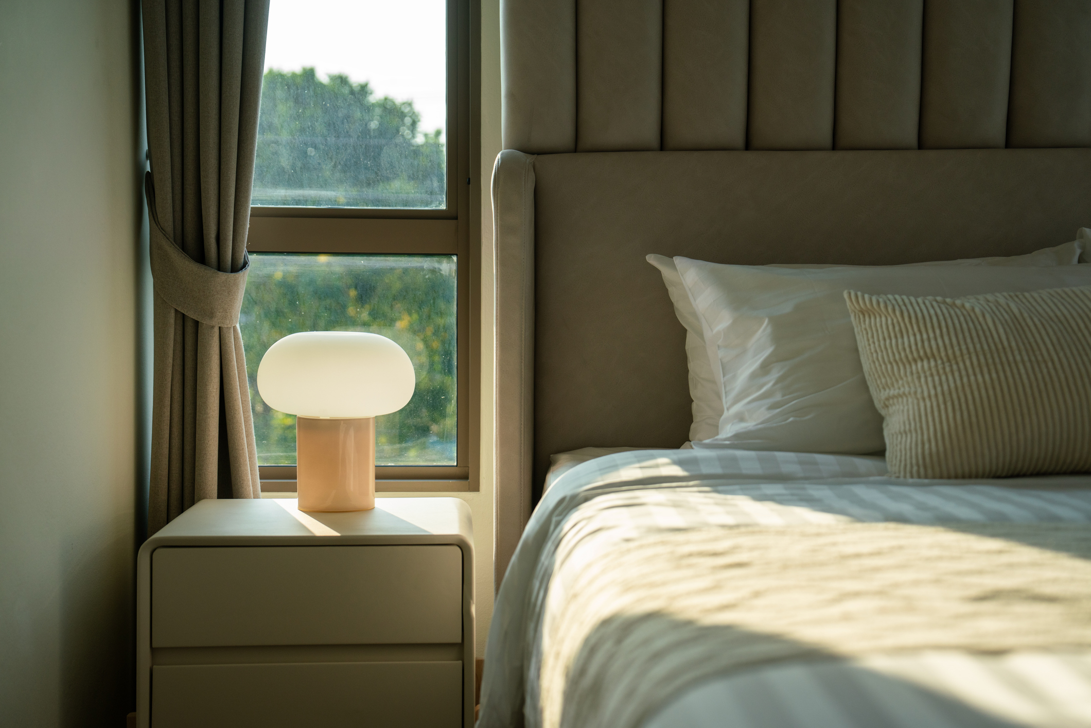 A warm and inviting bedroom featuring a neatly made bed, a stylish bedside table with a glowing lamp, and natural light filtering through the window.