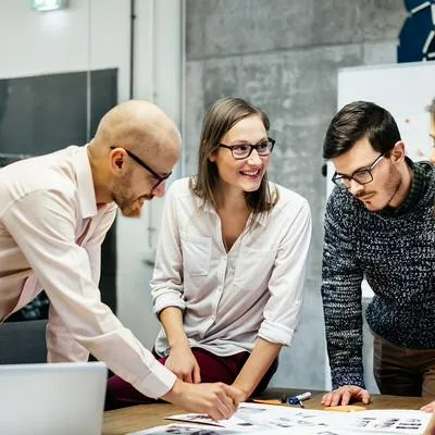 A team of four business people is standing in front a desk in a bright office room. They ambitiously point at documents while smiling and looking at the charts and notes.