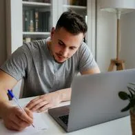 Hombre escribiendo en papel mientras está sentado con un computador portátil en su casa. En relación con cursos de Google.