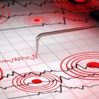 Close-up of a seismograph machine needle tracing seismic waves, indicative of earthquake activity, on graph paper with epicenter rings.