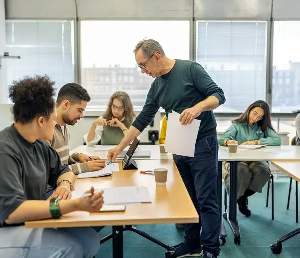 Senior professor talks with male student sitting at desk in adult education classroom. Male teacher showing something on a paper to student during lecture.
