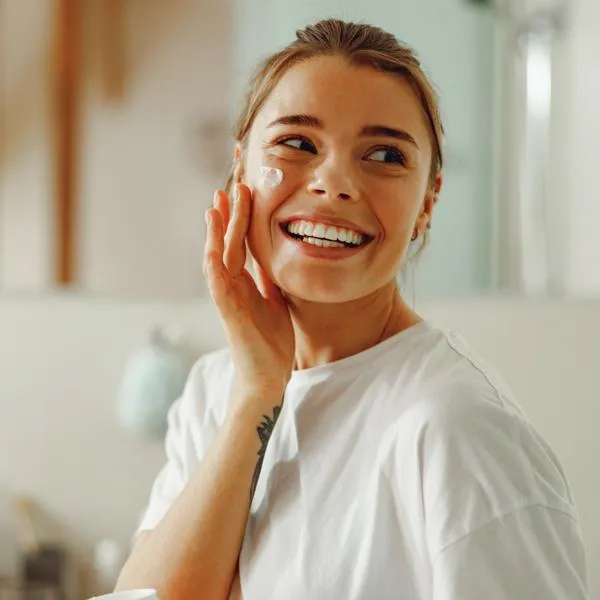 Happy woman applying hydrating moisturizer on her face sitting in bathroom. Home beauty routine