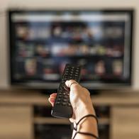 Close up of a person holding a control remote with a television screen on the background. A woman holding a remote control switches programs on the TV in the background.