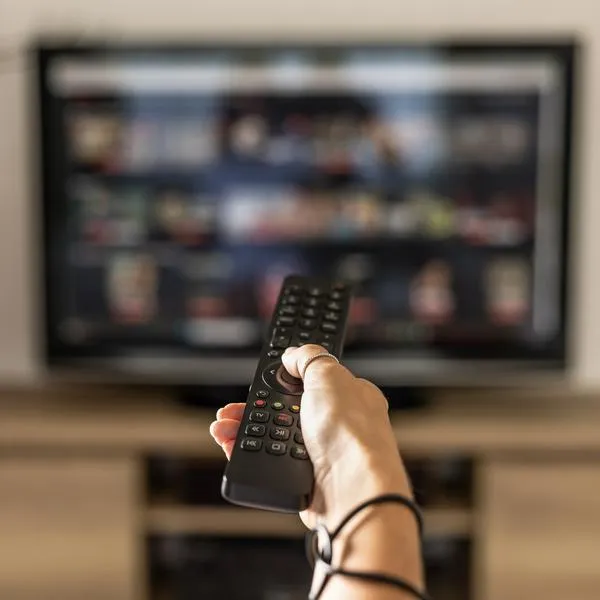 Close up of a person holding a control remote with a television screen on the background. A woman holding a remote control switches programs on the TV in the background.