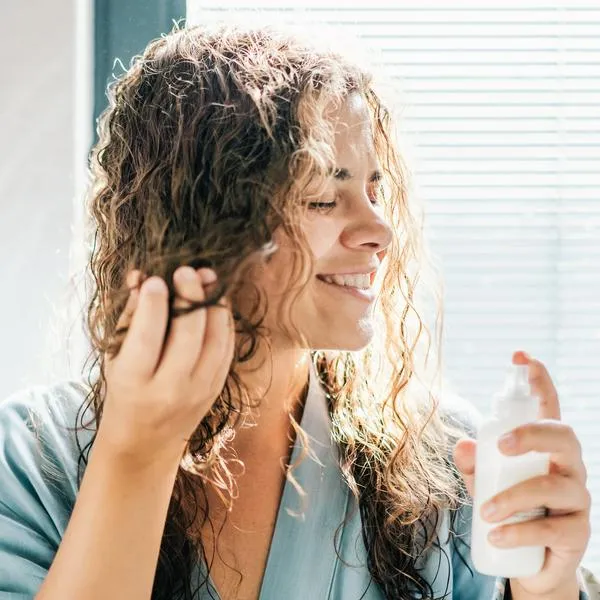 A woman with wet, wavy hair is applying a beauty product from a spray bottle. The bright backlighting creates a soft and intimate atmosphere, highlighting a self-care moment.
