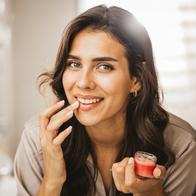 Happy young woman in pajama applying lip gloss in front of mirror in bathroom.