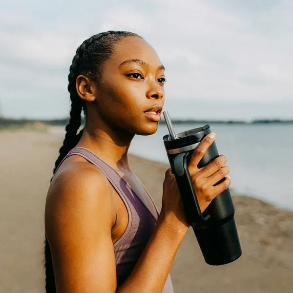 A young woman stands by a serene lakeside, sipping from a black tumbler while wearing athletic attire. The calm water and gentle sky create a tranquil background, enhancing the peaceful moment.