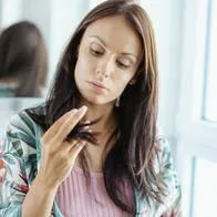 A woman looks down while brushing her long brown hair beside a mirror and window in a bright bathroom.