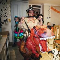 A front view of a young woman showing her funny side in her choice of inflatable Halloween costume which is her riding on the back of a dinosaur. You can also see different family members also in costume as they enjoy themselves at the family Halloween party!