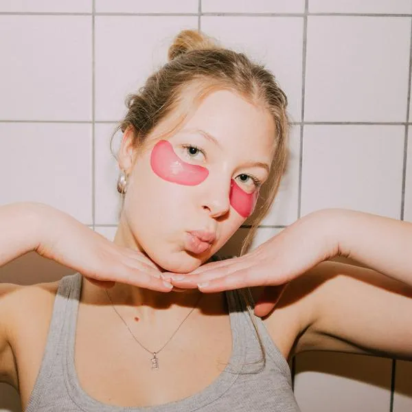 A young woman wears pink under-eye patches and poses with hands under her chin in a tiled bathroom. Casual tank top, skincare moment, fun vibe, bright lighting, and relaxed mood. Part of a series with video