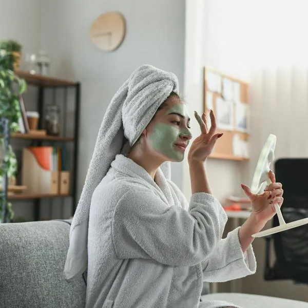 A woman in a plush bathrobe with a green facial mask and a towel wrapped on her head pamper herself at home, looking into a mirror for a calm, relaxing skincare routine.