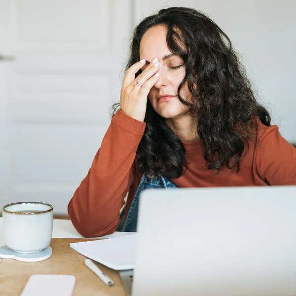Unhappy exhausted young woman feeling burned out while working late on her laptop at home. Depicts remote work stress, digital exhaustion and mental overload. Home office setting with warm lighting. Work-life balance challenges concept