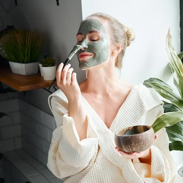 Positive young female in white bathrobe applying purifying clay mask on face with brush from bowl while sitting near window and relaxing during skincare procedure at home