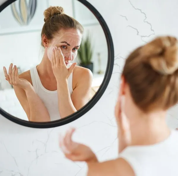 Young female reflected in mirror applying cleaning cosmetic mask on face during daily skincare routine in bathroom