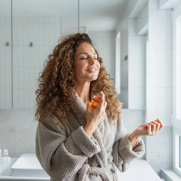 A mid adult woman with curly hair wearing a cozy robe applies fragrance in a bright modern bathroom with white tiles and natural daylight.