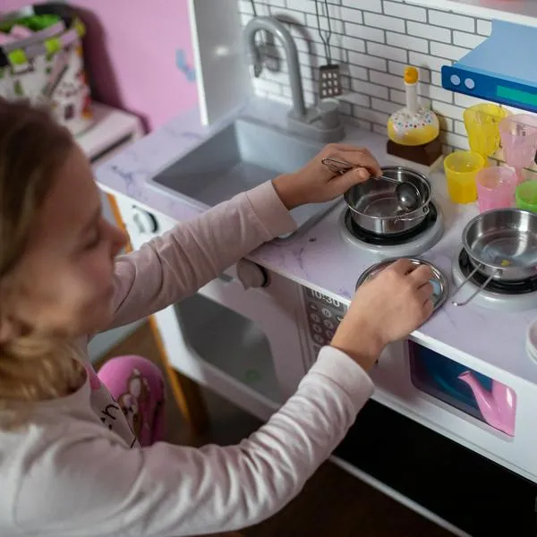 Smiling and engaged, a little girl role-plays as a chef, using her toy stove, pots, and colorful accessories. The play kitchen in her vibrant room offers a perfect space for learning and fun.