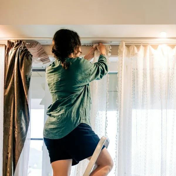 Woman standing on stepladder and hanging curtains in bedroom
