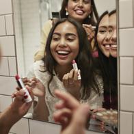 Smiling female friends applying lipstick while reflecting in mirror at home