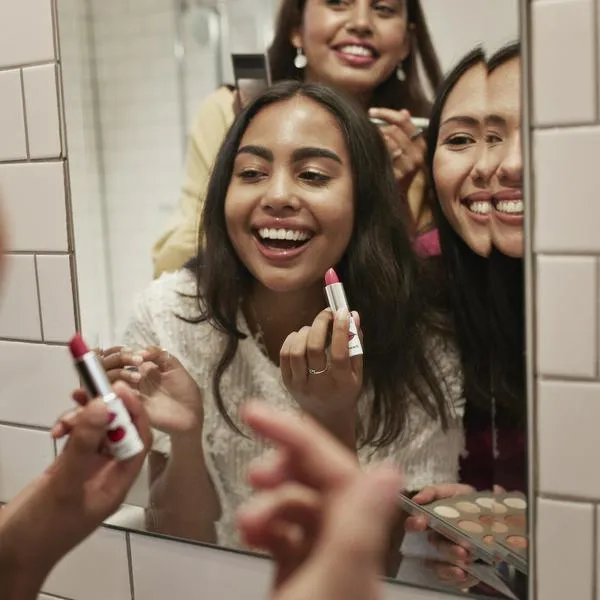 Smiling female friends applying lipstick while reflecting in mirror at home