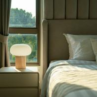 A warm and inviting bedroom featuring a neatly made bed, a stylish bedside table with a glowing lamp, and natural light filtering through the window.