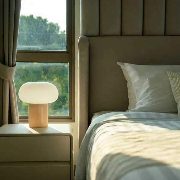 A warm and inviting bedroom featuring a neatly made bed, a stylish bedside table with a glowing lamp, and natural light filtering through the window.
